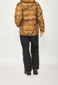 Braune gepolsterte Jacke mit marmoriertem Muster, kombiniert mit schwarzen Hosen. Verfügt über eine Kapuze und eine strukturierte Oberfläche. Schwarze Stiefel runden das Outfit ab.