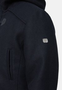 Dunkelblaue Kapuzenjacke aus strukturiertem Stoff, mit teilweisem Reißverschluss und einem kleinen Metall-Logo-Patch am Ärmel, ausgestattet mit schräg angeschnittenen Fronttaschen.