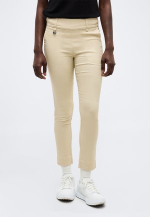 Persoon draagt beige, slimvallende enkellange broek, witte sokken, witte sneakers en een zwart topje, staand tegen een effen achtergrond.