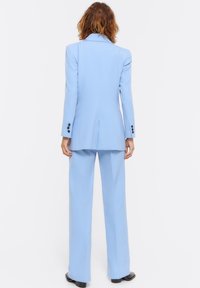 Person with shoulder-length brown hair wears a light blue tailored suit with black shoes, standing facing away on a white background.