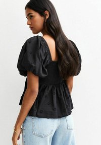 Black puff-sleeve top with a smocked bodice, open back, and floral embroidery. Cotton fabric, relaxed fit, paired with light denim jeans.