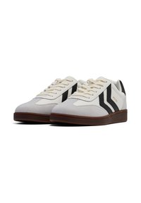 Sneakers bianchi e grigio chiaro a taglio basso con suole marroni e strisce nere laterali, mostrate da un'angolazione bassa su uno sfondo bianco.