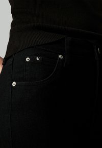 Pantalones vaqueros de denim negro con una forma ajustada, presentan un herraje plateado y un parche con el logo en el bolsillo delantero. Tejido texturizado con un acabado suave.