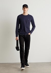 Μακρυμάνικο μπλουζάκι ναυτικού χρώματος, μαύρο παντελόνι και μαύρα αθλητικά παπούτσια. Το άτομο κρατά ακουστικά και φοράει ένα μαύρο καπέλο με λογότυπο.