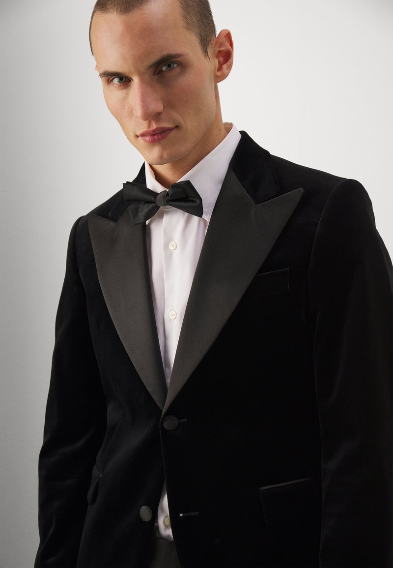 Czarna welwetowa marynarka smokingowa z satynowymi klapami w kształcie szpica, w zestawie z białą koszulą i czarną muszką. Posiada 1 kieszeń i akcenty w postaci guzików.