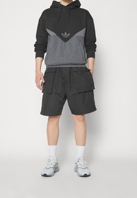 Person som bär en svart och grå huoodie, svarta cargoshorts med stora fickor, vita strumpor och vita sneakers med blå sulor.