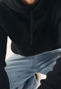 Svart stickad tröja med hög krage och framknapp, med en texturerad yta. Matchad med ljusblå denimjeans.