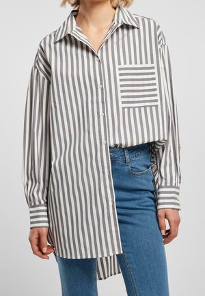 Femme portant une chemise blanche oversize à rayures grises verticales et une poche rayée horizontale contrastante, associée à un jean bleu.