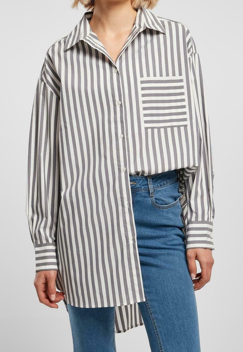 Femme portant une chemise blanche oversize à rayures grises verticales et une poche rayée horizontale contrastante, associée à un jean bleu.