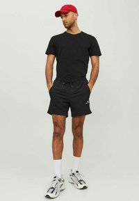 Schwarzes T-Shirt und schwarze Shorts aus leichtem Material, kombiniert mit einer roten Mütze und weißen Sportsocken. Das Schuhwerk hat eine Mischung aus Farben.