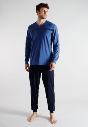 Junger Mann steht barfuß, trägt ein blaues Langarmhemd und dunkle Hose, schaut nach links vor einfachem weißem Hintergrund.