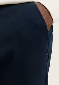 Pantalones deportivos azul marino con una textura suave, que presentan un bolsillo lateral y una etiqueta de logo cosida. El diseño incluye piernas ajustadas y una cintura elástica.