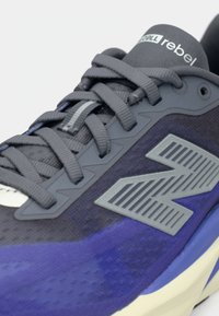 Αθλητικό παπούτσι σε γκρι και βαθύ μωβ χρώμα με υφασμάτινη υφή, χαρακτηριστικά κορδόνια και μεγάλο γκρι λογότυπο της μάρκας με ριγέ λεπτομέρειες στο πλάι.