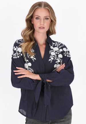 Kobieta o falowanych blond włosach nosi granatową bluzkę z białą kwiatową haftem i zawiązywanymi mankietami, stojąc z założonymi ramionami.