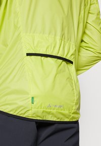 Lengvas, ryškiai laimo žalios spalvos švarkas su lygiu paviršiumi, turintis juodo apvado ir Vaude logotipo apatiniame krašte bei užtrauktuko kišene.
