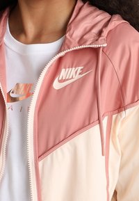 Person som bär en rosa och cremefärgad Nike vindjacka över en vit Nike Air T-shirt med logotyper synliga på båda plaggen.
