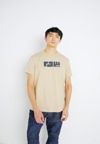 Beige katoenen t-shirt met een zwart grafisch ontwerp op de voorkant. Korte mouwen en een ronde halsontwerp. Draag het met donkerblauwe denim jeans.
