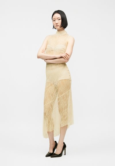 Γυναίκα με κοντά μαύρα μαλλιά στέκεται με σταυρωμένα τα χέρια, φορώντας ένα διάφανο μπεζ φόρεμα από δαντέλα πάνω από γυμνά εσώρουχα και μαύρες ψηλοτάκουνες γόβες.