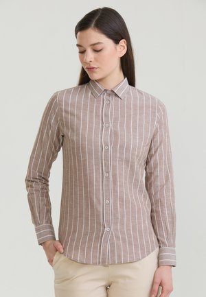 Kobieta nosząca beżową koszulę na długi rękaw z białymi pionowymi paskami, stojąca z jedną ręką w kieszeni beżowych spodni.