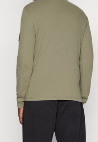 Chemise à manches longues vert olive en tissu ribbed texturé, col rond, avec un accent noir sur la manche gauche. Associée à un pantalon noir.