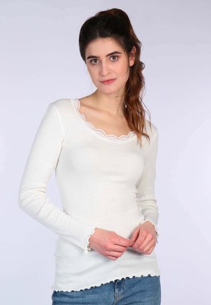 Weißes Langarmshirt mit geripptem Stoff, verziert mit einem gewellten Spitzenkragen und Bündchen. Kombiniert mit Jeans.