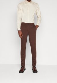 Brown tailored trousers made of a smooth fabric, featuring a slim fit, front pockets, and a button closure. Worn with a light-colored shirt.