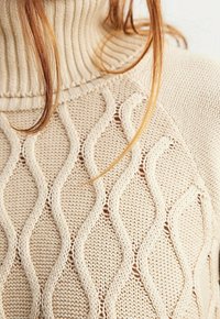 Beżowy sweter wykonany na drutach z prążkowanym golfem oraz falistą strukturą z otwartymi przeszyćmi, tworzącą lekki, warstwowy efekt.