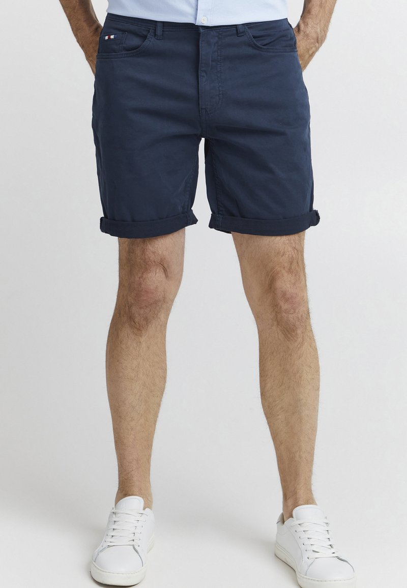 Shorts din bumbac bleu marin, cu manșete răsucite, având buzunare frontale și cusături contrastante. Purtați cu adidași albi.