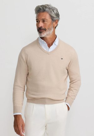 Uomo di mezza età con capelli grigi e barba che indossa un maglione beige a scollo a V sopra una camicia bianca con colletto e pantaloni bianchi, in piedi contro uno sfondo semplice.