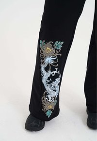Zwarte broek met gedetailleerde borduursels van een blauwe draak en bloemmotieven in groen en goud langs de zijkant. Geweven stof met een casual pasvorm.
