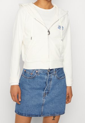 Personne portant un sweat à capuche blanc zippé avec un petit logo et une jupe en jean bleue, debout devant un fond blanc uni.