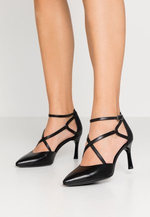 Czarne szpilki mają spiczasty nos, pasek wokół kostki z krzyżującym się wzorem oraz gładką skórzaną teksturę.