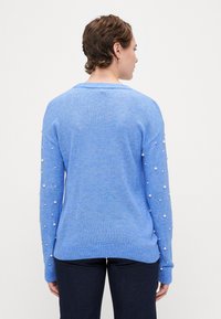 Lyseblå genser med ribbet halslinning og mansjetter, med perle-detaljer langs ermene. Myk tekstur og avslappet passform.