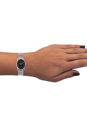 Montre-bracelet en maille argentée avec un cadran rond noir, comportant des index et des aiguilles argentés, portée sur une main avec du vernis à ongles noir.