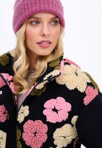 Chlpatá čierna bunda s farebnými kvetinovými vzormi v ružovej, krémovej a olivovej farbe. Skombinovaná s rebrovým ružovým čiapkom. Detailný portrét.