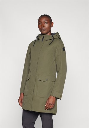 Person, der eine grüne Kapuzenparka mit Vordertaschen und schwarzen Kordeln trägt, steht vor einem schlichten weißen Hintergrund.