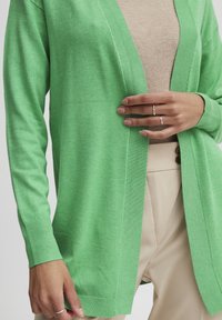 Osoba w zielonym kardiganie, nałożonym na beżową bluzkę i beżowe spodnie, prezentująca srebrne pierścionki na palcach.