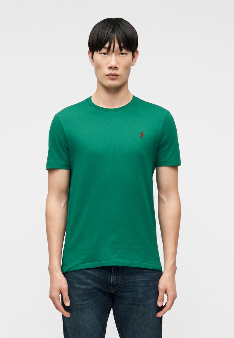 Νεαρός άνδρας με ίσια μαύρα μαλλιά που φοράει πράσινο κοντομάνικο μπλουζάκι και σκούρο τζιν, στέκεται μπροστά σε απλό λευκό φόντο.