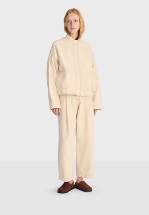 Femme debout portant une veste et un pantalon oversize beige avec des chaussons marron et des chaussettes rayées sur un fond clair uni.