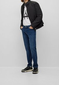 Svart bomberjacka, vit t-shirt med tryck, blå jeans och svarta sneakers med beige accenter. En avslappnad outfit som kontrasterar färger och texturer.