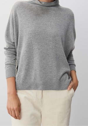 Femme portant un pull ample gris clair à col haut et un pantalon en velours côtelé crème, debout avec une main dans la poche.