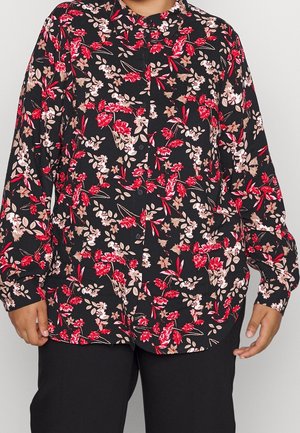 Persoon draagt een zwart overhemd met knopen met rode, roze en beige bloemmotieven en zwarte broek, staand met armen ontspannen langs het lichaam.