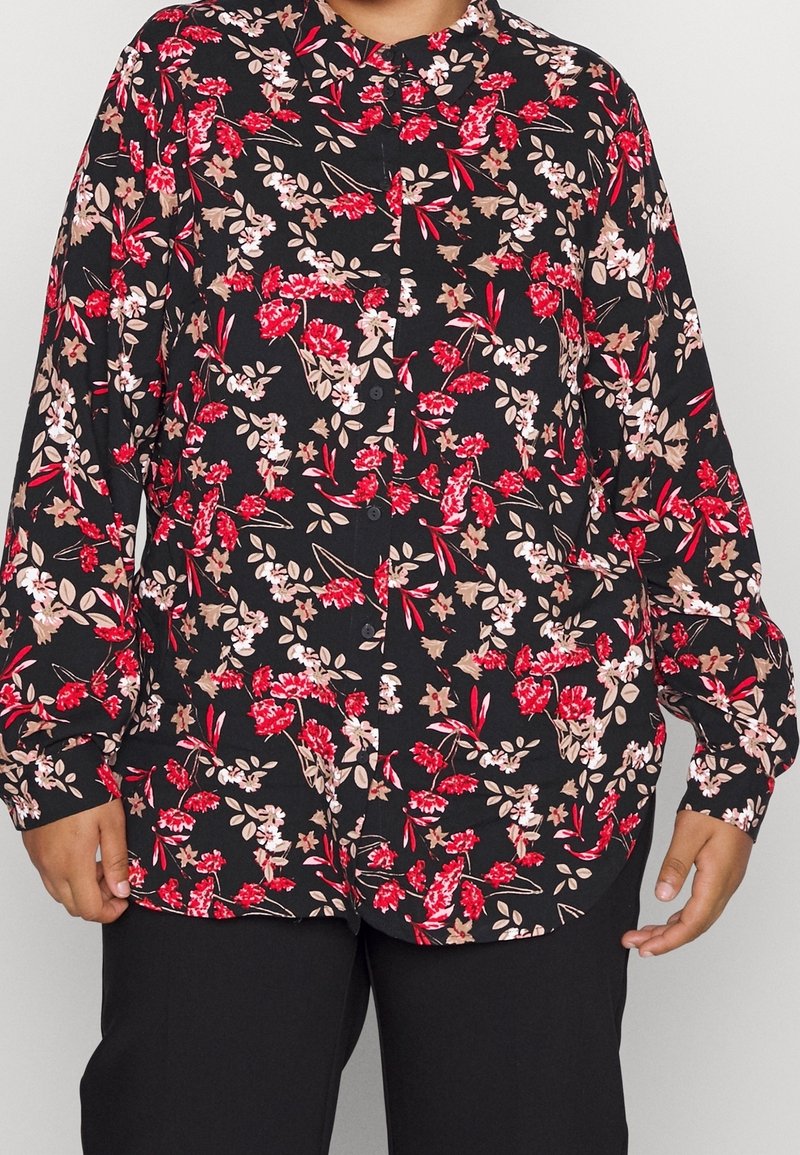 Personne portant une chemise noire boutonnée avec des motifs floraux rouges, roses et beiges et un pantalon noir, debout les bras détendus le long du corps.