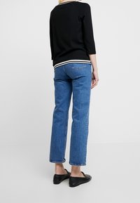 Persoon in blauwe rechte jeans, zwarte instapschoenen en een zwart shirt met witte strepen aan de onderkant, staande tegen een simpele witte achtergrond.