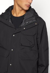 Giacca nera esterna in nylon, con zip frontale, bottoni a pressione, due tasche frontali, cappuccio e una fodera liscia e strutturata.
