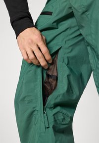 Žalios neperšlampamos kelnės su užtrauktuko kišene, matoma tinklelio pamušalu ir siūlių detalėmis. Ranka atidaro šoninį užtrauktuką.