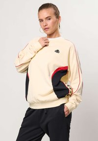 Žena v krémovej mikine s červenými a čiernymi detailmi a logom Kappa, v čiernych nohaviciach, pózujúca s jednou rukou pri golieri.