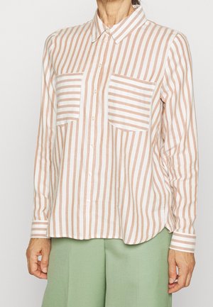 Kobieta ubrana w beżową koszulę zapinaną na guziki z pionowymi biało-beżowymi paskami oraz poziomymi paskami na kieszeniach piersiowych, a także zielone spodnie.