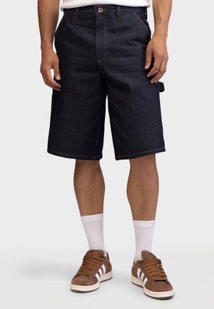 Mężczyzna ubrany w ciemne, dżinsowe spodenki do kolan, białe skarpetki do kostek oraz brązowe zamszowe trampki z białymi paskami, stojący na tle jednolitego tła.