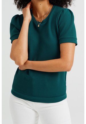 Teal struktureret top med rund hals og korte ærmer; har en tætsiddende silhuet og en glat, ensfarvet kant. Båret med hvide bukser.